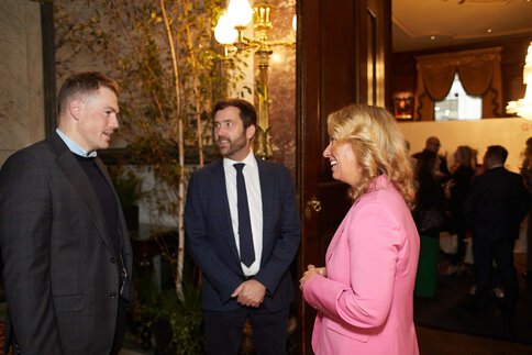 Zwei Männner und eine Frau in Businesskleidung stehen gemeinsam vor dem Eingang zu einem gut besuchten Saal und unterhalten sich.