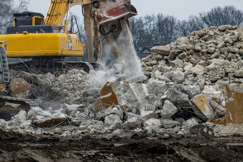 Excavator with hydraulic hammer breaking concrete rubble on a demolition site, surrounded by debris and exposed reinforcing steel.