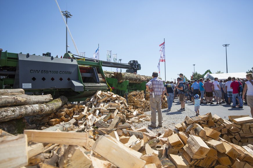 Visitors to INTERFORST observe a large green automatic splitting machine on the outdoor exhibition grounds at Messe München. Piles of split timber lie next to it.