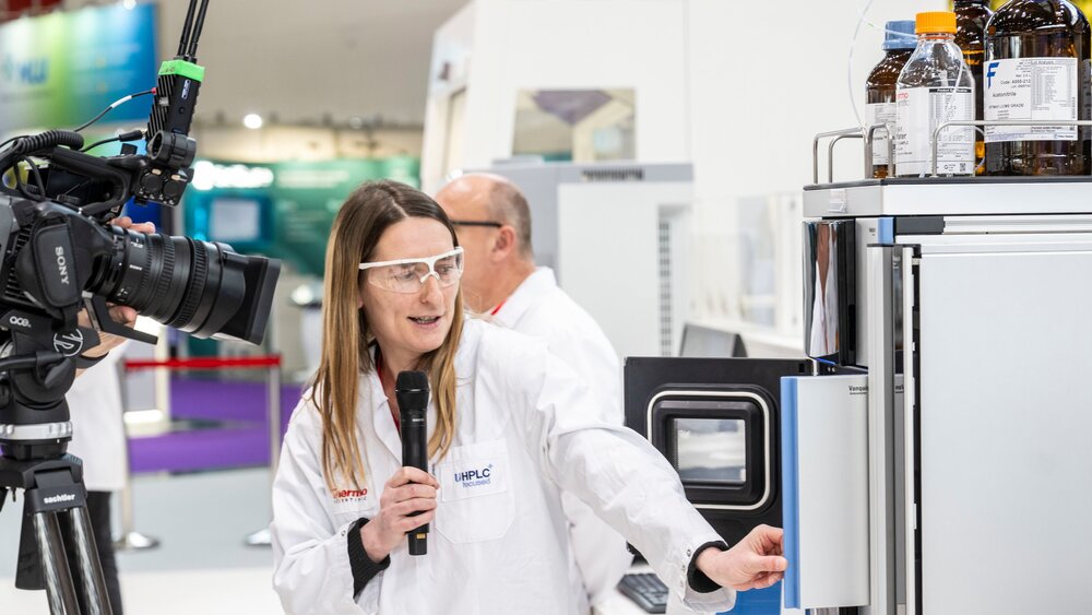 A person in a lab coat explains an analytical instrument on camera and demonstrates how to use it with a microphone at a trade show.