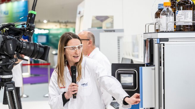 A person in a lab coat explains an analytical instrument on camera and demonstrates how to use it with a microphone at a trade show.