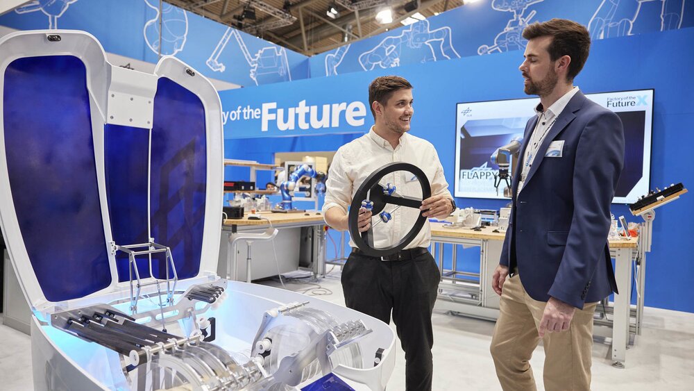 Two men discussing at a trade fair stand next to an unfolded, futuristic-looking machine.