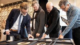Vier Männer begutachten Schmuckstücke auf Ausstellungstischen in einem Ausstellungsraum.