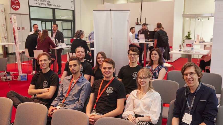 A group of people are sitting on chairs during a lecture, with some participants standing in the background.