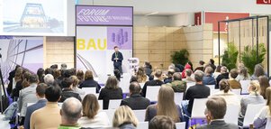 Vortragssituation auf einer Messe: Ein Referent steht auf einer Bühne mit der Aufschrift „Forum Future of Building / BAU“ vor einer Leinwand mit Gebäudeabbildung, das Publikum sitzt in Reihen und hört zu.