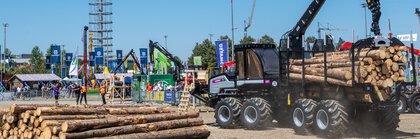 Forstmaschine lädt Baumstämme auf einem Vorführgelände, während Besucher eine Demonstration auf einer Fachmesse beobachten.