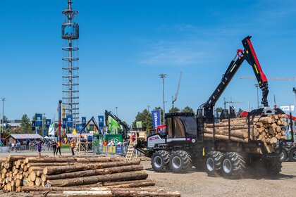 Forstmaschine lädt Baumstämme auf einem Vorführgelände, während Besucher eine Demonstration auf einer Fachmesse beobachten.