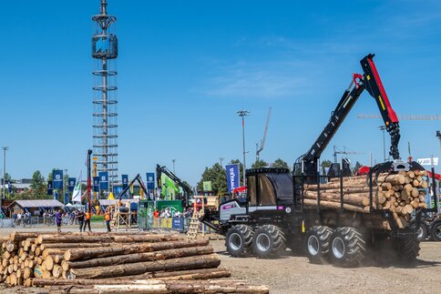 Forestry machine loading logs at a demonstration area while visitors watch at a trade fair.