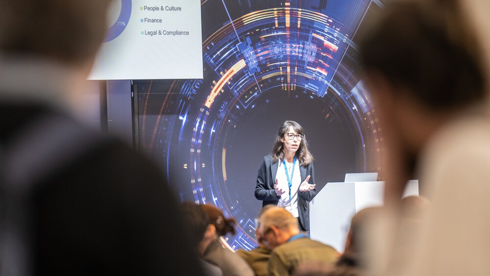 A woman gives a presentation to an audience on a stage with a digital background.