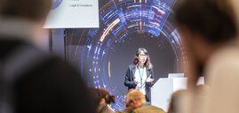 Eine Frau hält vor einem Publikum auf einer Bühne mit digitalem Hintergrund eine Präsentation.