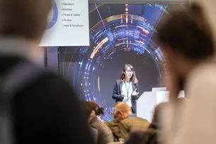 A woman gives a presentation to an audience on a stage with a digital background.