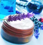 A sprig of lavender lies on an open glass cream jar, surrounded by shades of blue and lavender flowers