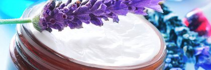 A sprig of lavender lies on an open glass cream jar, surrounded by shades of blue and lavender flowers