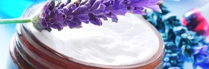 Ein Lavendelzweig liegt auf einem offenen Cremetöpfchen aus Glas, umgeben von Blautönen und Lavendelblüten