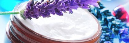 Ein Lavendelzweig liegt auf einem offenen Cremetöpfchen aus Glas, umgeben von Blautönen und Lavendelblüten
