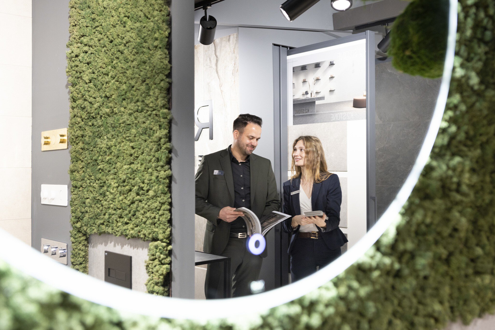 View into a round mirror in which a man and a woman can be seen talking with brochures in their hands at a trade fair stand at BAU.
