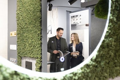 Blick in einen runden Spiegel, in dem ein Mann und eine Frau zu sehen sind, die sich mit Prospekten in der Hand an einem Messestand auf der BAU unterhalten.