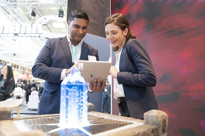 Ein Mann und eine Frau in Businesskleidung stehen hinter einem Steinpodest mit einer Art blau leuchtenden Kristall und blicken gemeinsam auf ein Tablet.