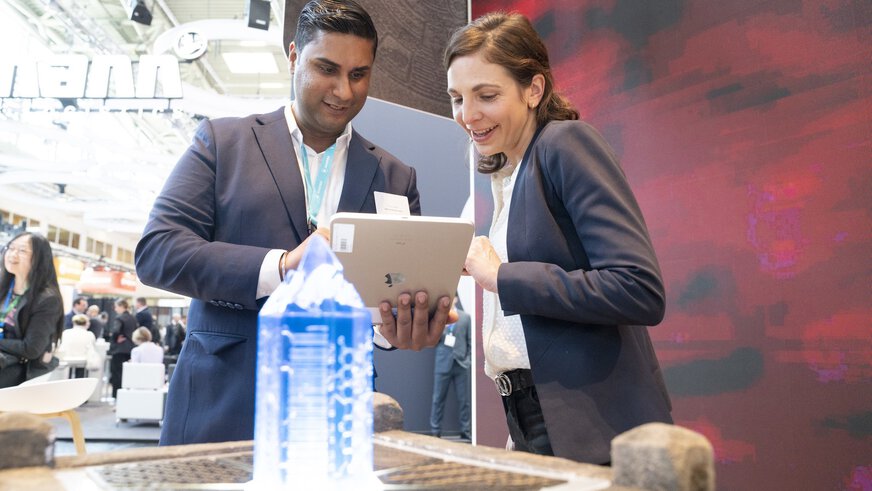 Ein Mann und eine Frau in Businesskleidung stehen hinter einem Steinpodest mit einer Art blau leuchtenden Kristall und blicken gemeinsam auf ein Tablet.
