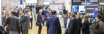 Numerous trade fair visitors stand in groups between two rows of exhibition stands at LOPEC and chat.