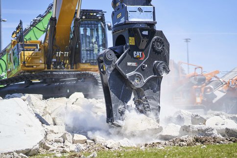 Excavator with hydraulic attachment crushes chunks of concrete under a blue sky.