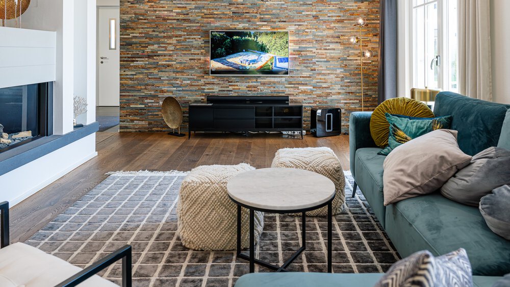 Modernes Wohnzimmer mit Steinakzentwand, Fernseher, blauem Sofa und runden Couchtischen auf einem gemusterten Teppich.