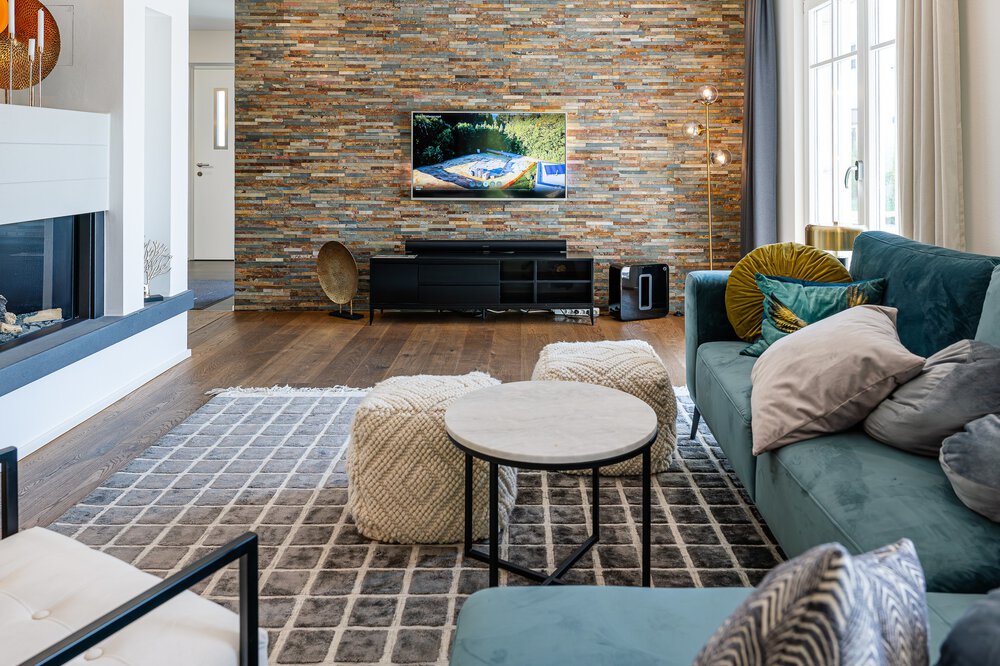 Modernes Wohnzimmer mit Steinakzentwand, Fernseher, blauem Sofa und runden Couchtischen auf einem gemusterten Teppich.