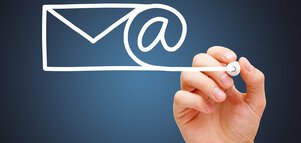 Eine Hand zeichnet mit einem weißen Marker das Symbol eines Briefumschlags, das mit einem @-Symbol verbunden ist in die Luft.