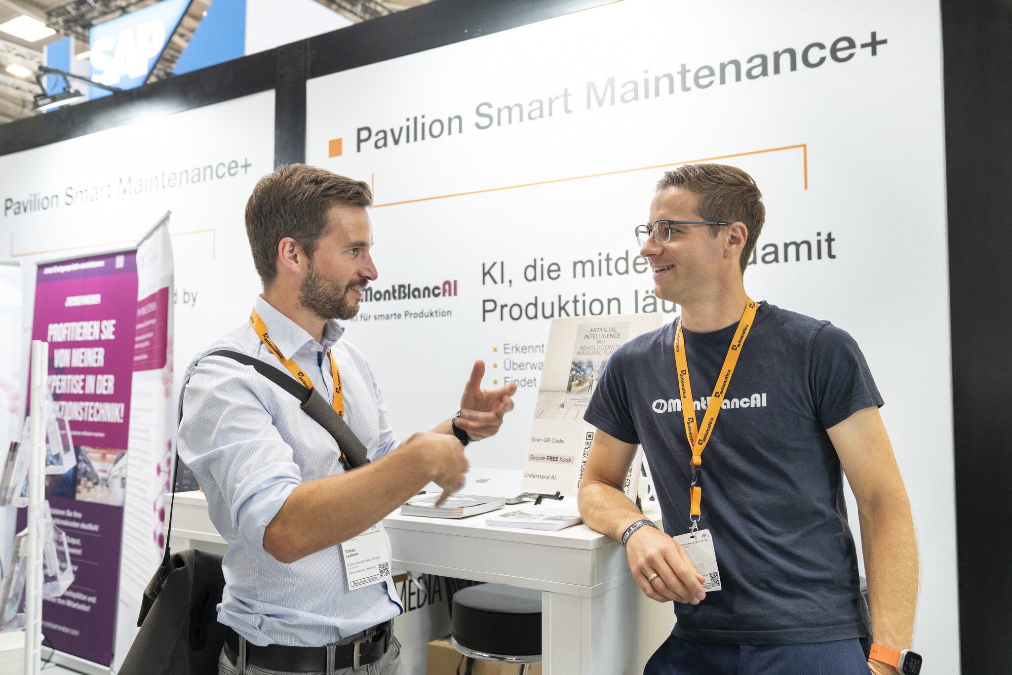 Zwei Männer mit Konferenzschildern unterhalten sich an einem Stand mit der Aufschrift Pavilion Smart Maintenance+ auf einer Veranstaltung.