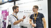 Zwei Männer mit Konferenzschildern unterhalten sich an einem Stand mit der Aufschrift Pavilion Smart Maintenance+ auf einer Veranstaltung.