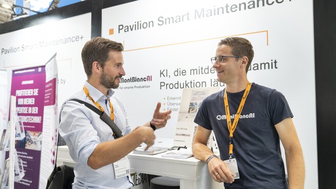 Zwei Männer mit Konferenzschildern unterhalten sich an einem Stand mit der Aufschrift Pavilion Smart Maintenance+ auf einer Veranstaltung.