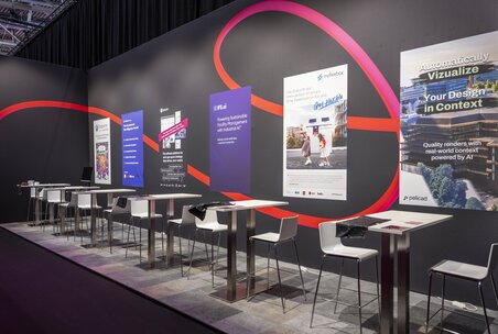 Eine Reihe von einzelnen Tischen mit jeweils zwei Stühlen mit zahlreichen Werbeplakaten dahinter in einer Ecke der EXPO REAL.