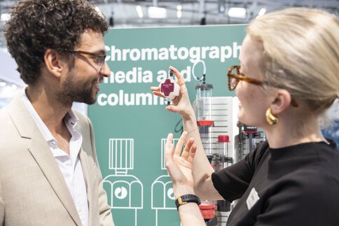 Zwei Personen an einem Messestand im Gespräch. Eine hält ein technisches Teil in der Hand. Im Hintergrund ist der Schriftzug 