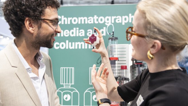 Two people talking at a trade fair stand. One is holding a technical part. The word 