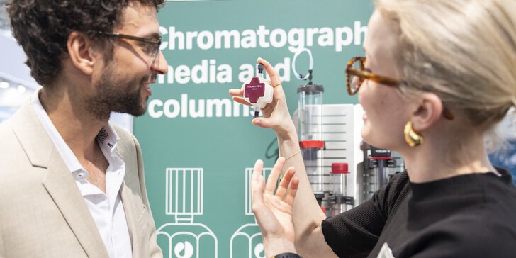 Zwei Personen an einem Messestand im Gespräch. Eine hält ein technisches Teil in der Hand. Im Hintergrund ist der Schriftzug 