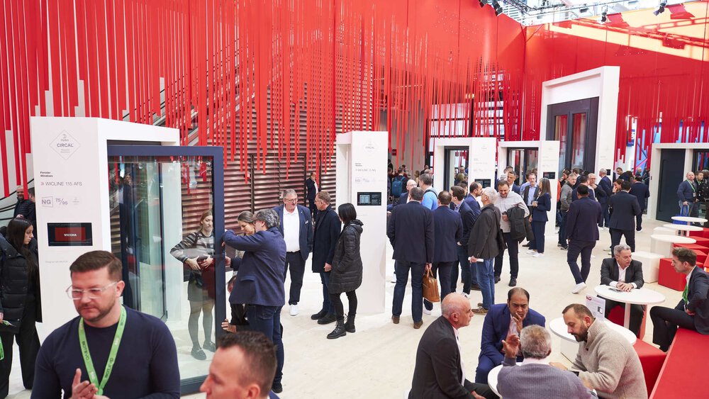 Many visitors in an exhibition room at BAU 2025, some looking at exhibits, others sitting together at round tables. Many red ribbons hang from the ceiling as decoration.