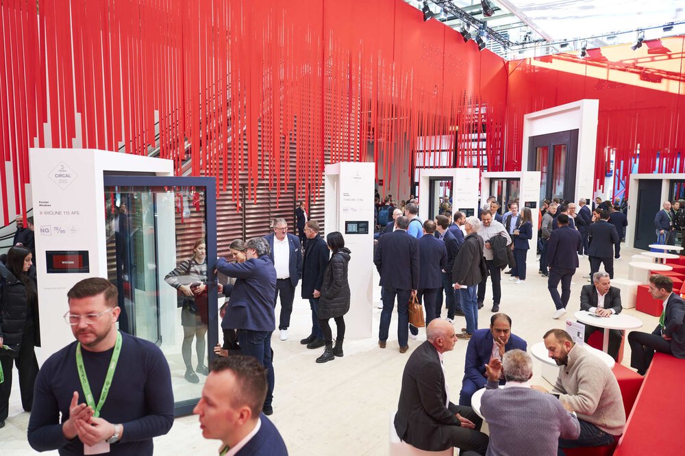 Viele Besucher in einem Ausstellungsraum der BAU 2025. Einige betrachten Ausstellungsstücke, andere sitzen zusammen an runden Tischen. Von der Decke hängen viele rote Bänder als Dekoration.