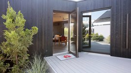 Eine offene Glastür am schwarzen Holzhaus führt zu einem modernen Innenraum mit Pflanzen und einer Willkommensmatte auf der Terrasse.