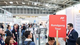 Guided trade fair tour with a group of visitors and a “Tour” sign.