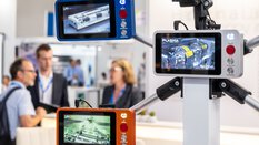 Three mounted screens display images of machines at a technology exhibition, with people in the background.