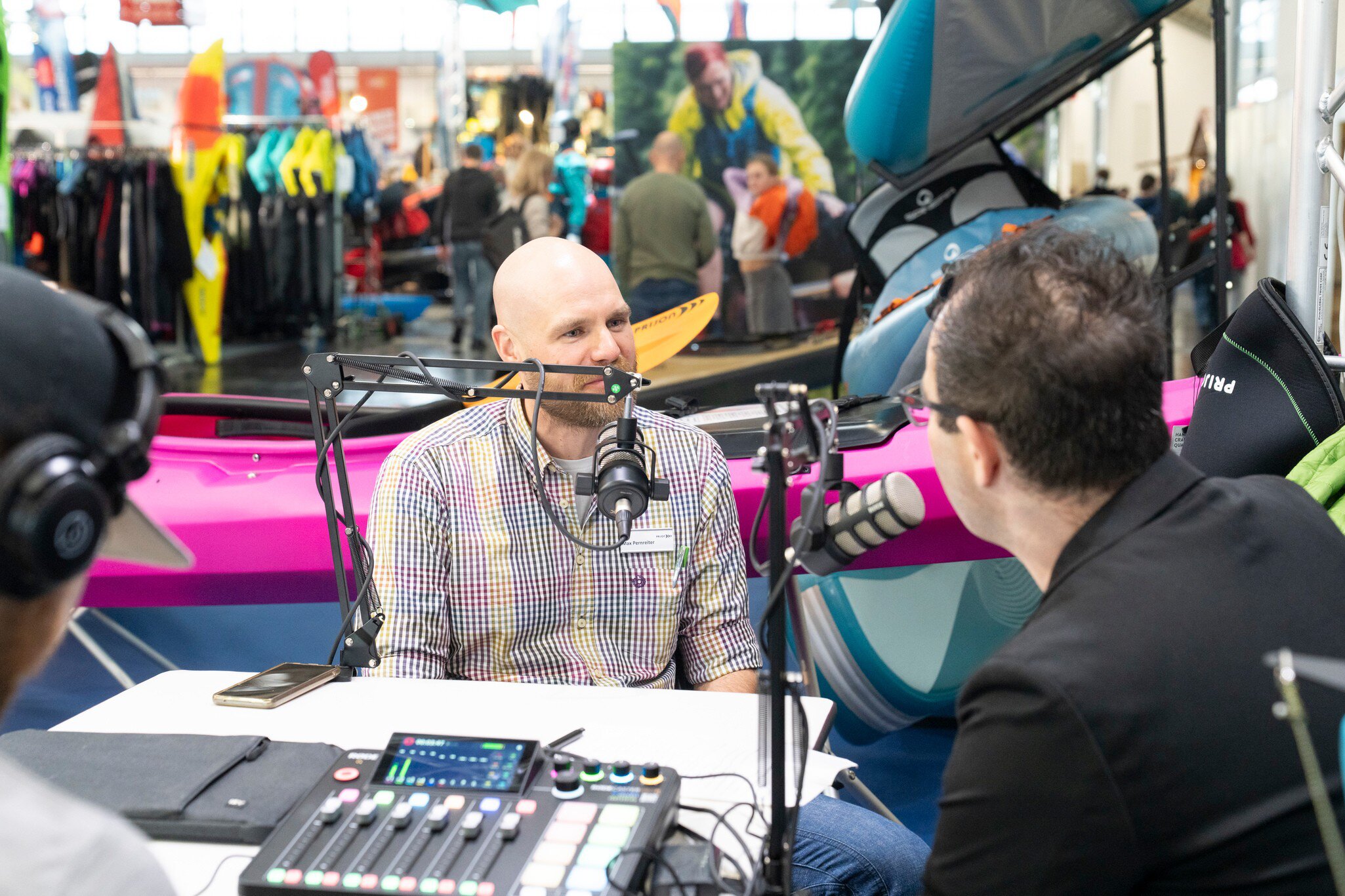 Drei Personen sitzen an einem Tisch mit Mikrofonen und Audio‑Mischpult, diskutieren vor einem pinken Kajak auf einer belebten Messe mit Ausstellungsständen.
