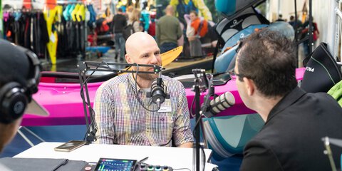 Drei Personen sitzen an einem Tisch mit Mikrofonen und Audio‑Mischpult, diskutieren vor einem pinken Kajak auf einer belebten Messe mit Ausstellungsständen.