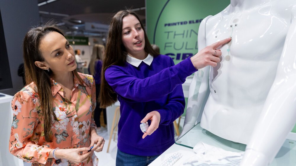 Two women are standing at a stand at LOPEC, looking at small medical components that are stuck to the torso of a white doll.