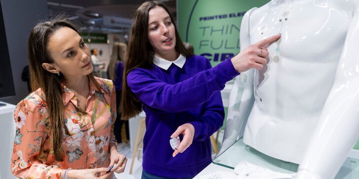 Two women are standing at a stand at LOPEC, looking at small medical components that are stuck to the torso of a white doll.
