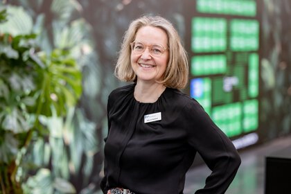 Portrait of a smiling woman wearing glasses and a black blouse with a visible name badge; she stands before a blurred leafy exhibition backdrop with potted plants behind her.