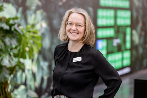Portrait of a smiling woman wearing glasses and a black blouse with a visible name badge; she stands before a blurred leafy exhibition backdrop with potted plants behind her.