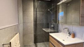 Modernes Badezimmer mit großen grauen Fliesen, einer begehbaren Dusche, einem hölzernen Waschtisch und Handtüchern auf einem Ständer.