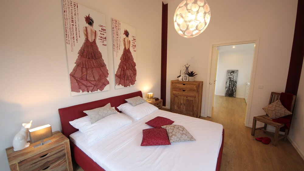 Blick in ein Schlafzimmer mit rotem Doppelbett, einer modischen runden Lampe, Beistelltischen und zwei großen Gemälden.