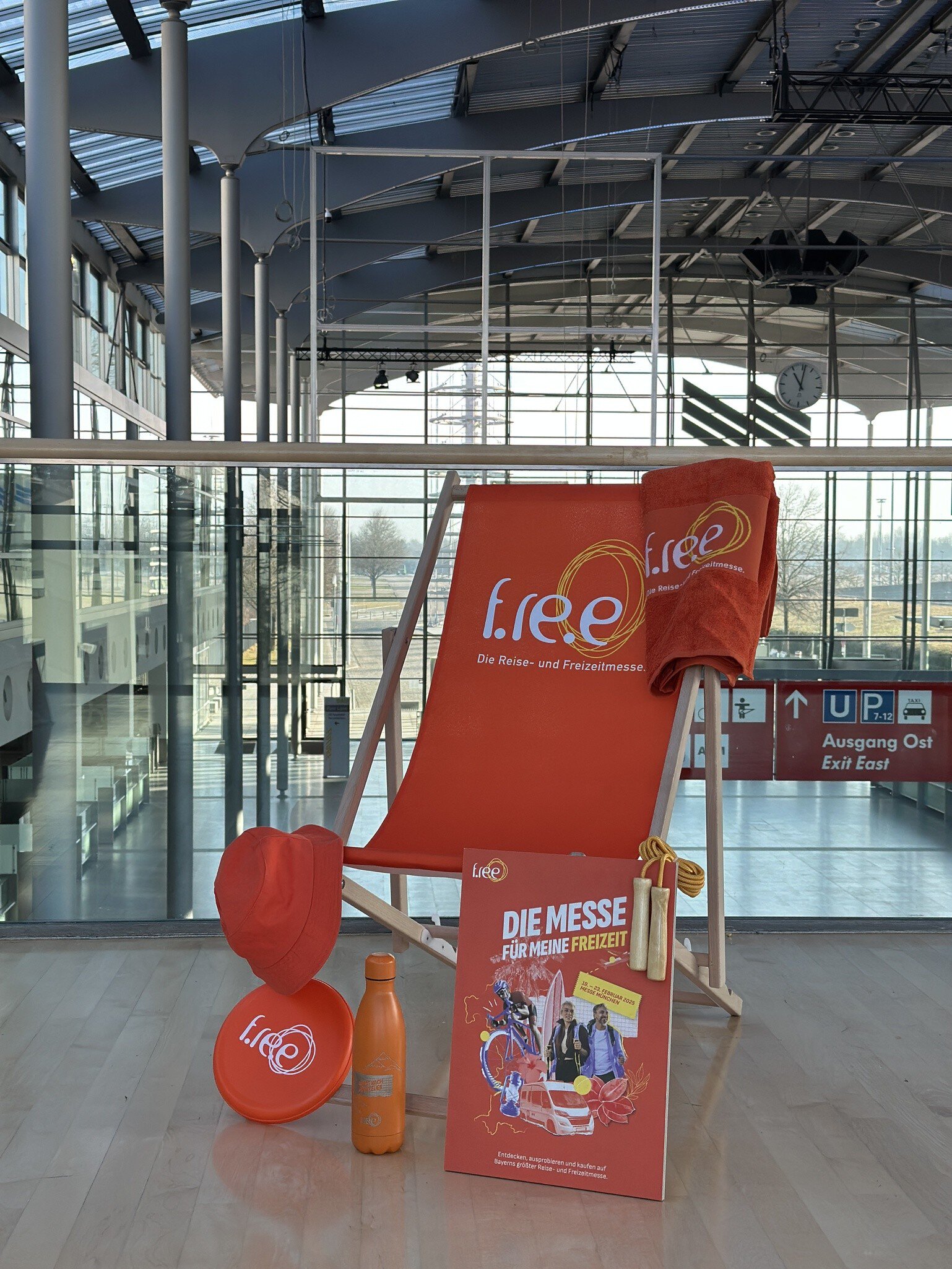 Ein orangener Liegestuhl der f.re.e Messe mit mehreren orangenen Artikel zur f.re.e: Eine Anglermütze, eine Frisbee, eine Trinkflasche und ein Badetuch und ein Plakat.