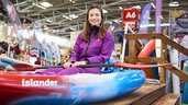 Lächelnde Frau im Kajak auf dem Indoor-Paddelbecken der Messe f.re.e, mit Paddel in der Hand. Es sind verschiedene Messestände im Hintergrund.